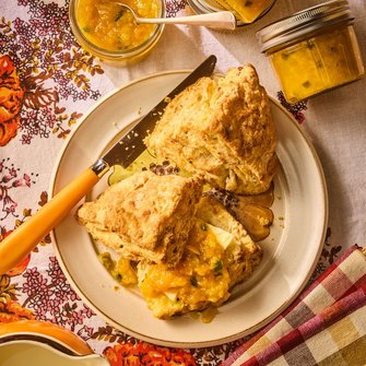 Fresh homemade scones with butter and peach jam on a rustic plate, surrounded by jars of fruit preserves.